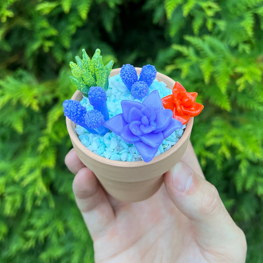 Hand holding a small pot with colorful miniature glass plants against a green foliage background.