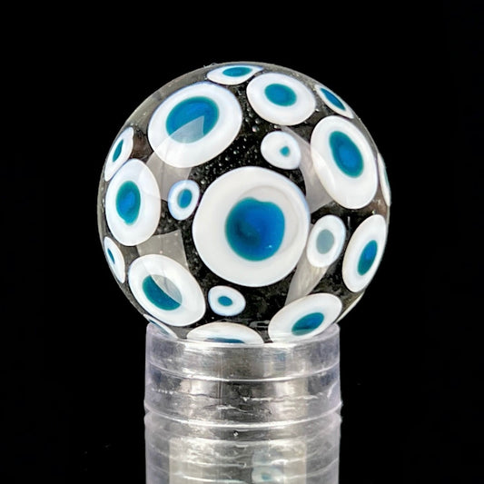 A glass marble with a design of blue dots on a white background, approximately 26mm in diameter, displayed on a clear stand.