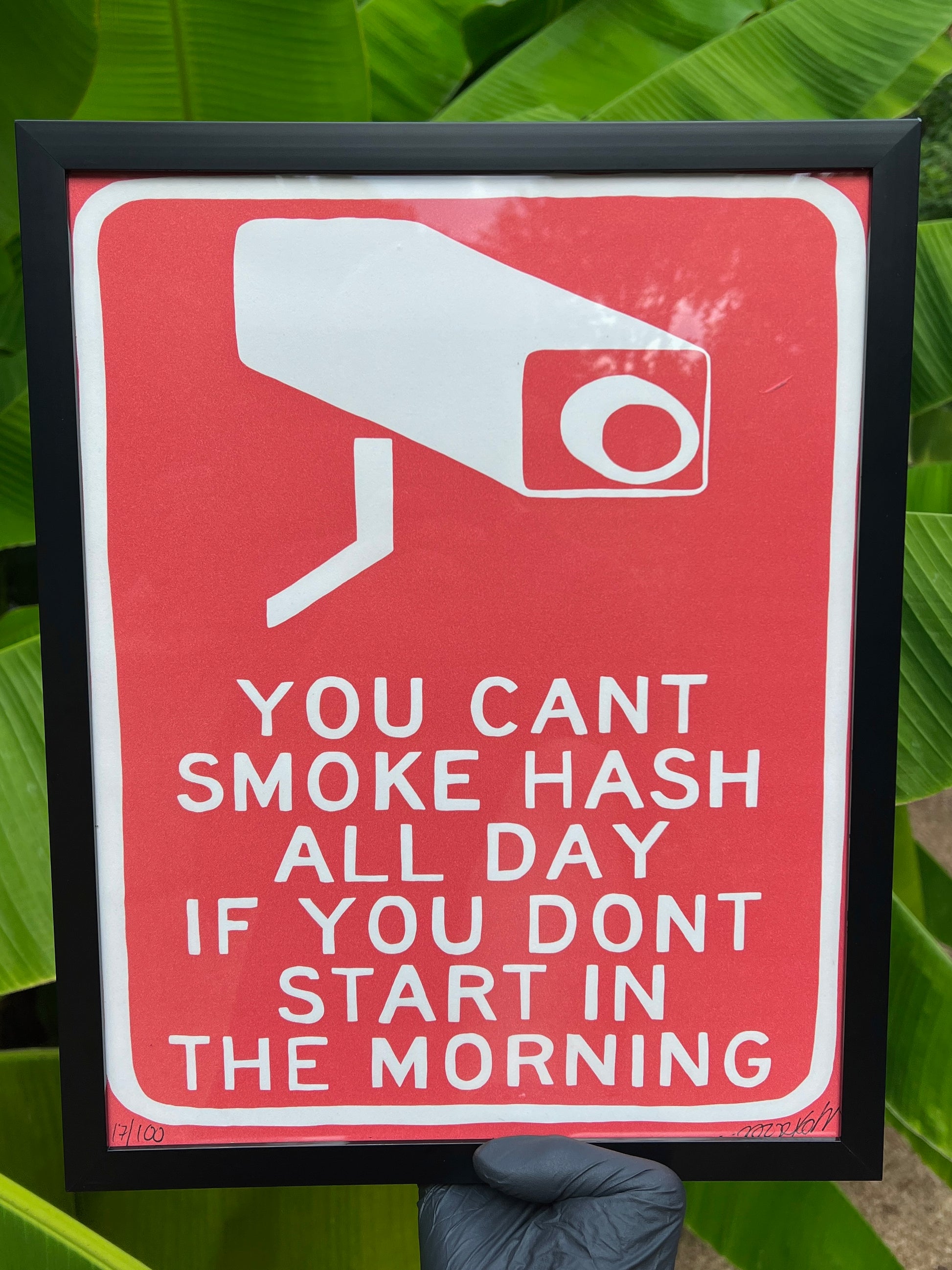 A red print with white text and a graphic of a security camera that says 'YOU CAN'T SMOKE HASH ALL DAY IF YOU DON'T START IN THE MORNING'.