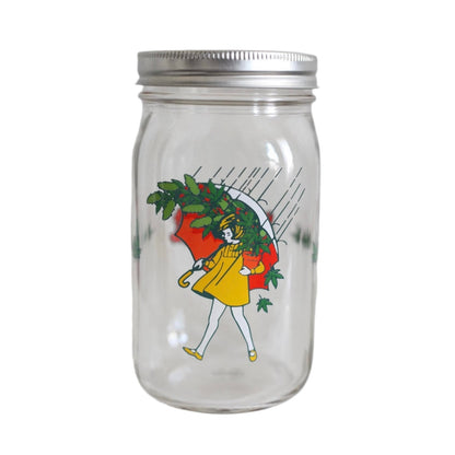 A clear glass ball jar with a illustration of a person with a green umbrella and leaves on the lid.