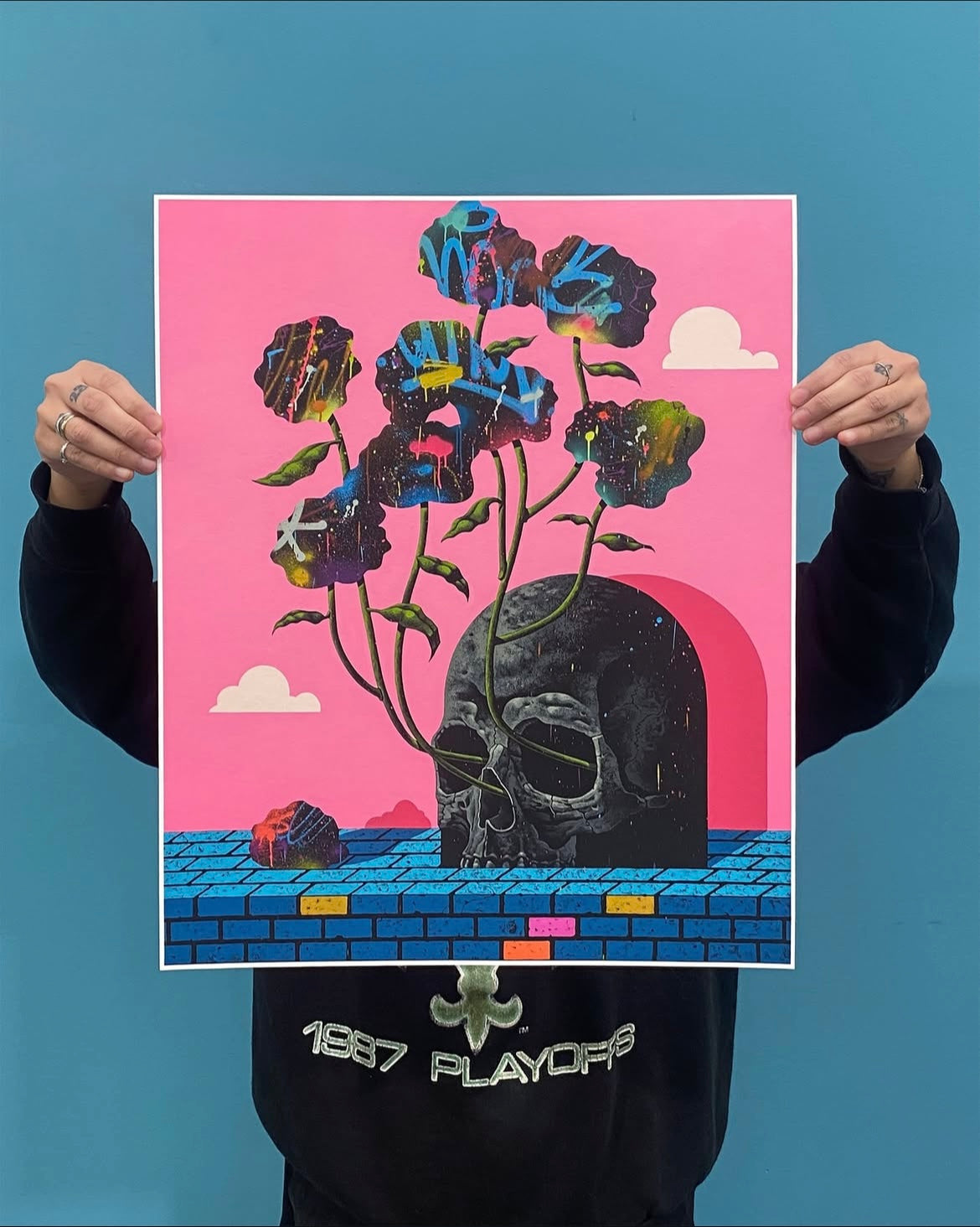 Person holding a colorful poster with a skull and flowers against a blue background.