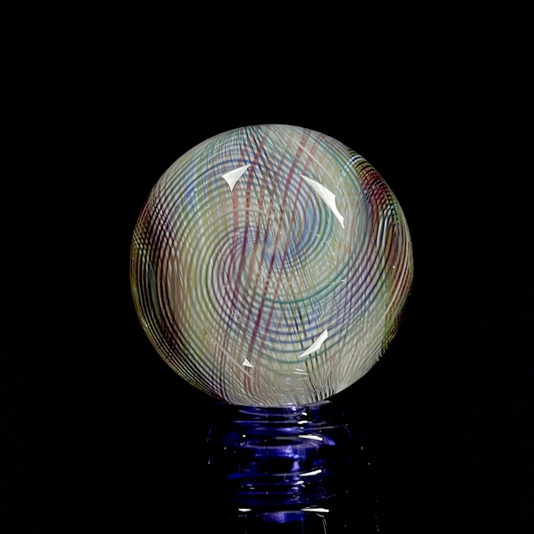 A multicolored glass marble with a mix of vibrant and muted tones, featuring a patterned design. The marble appears to have a convex surface.