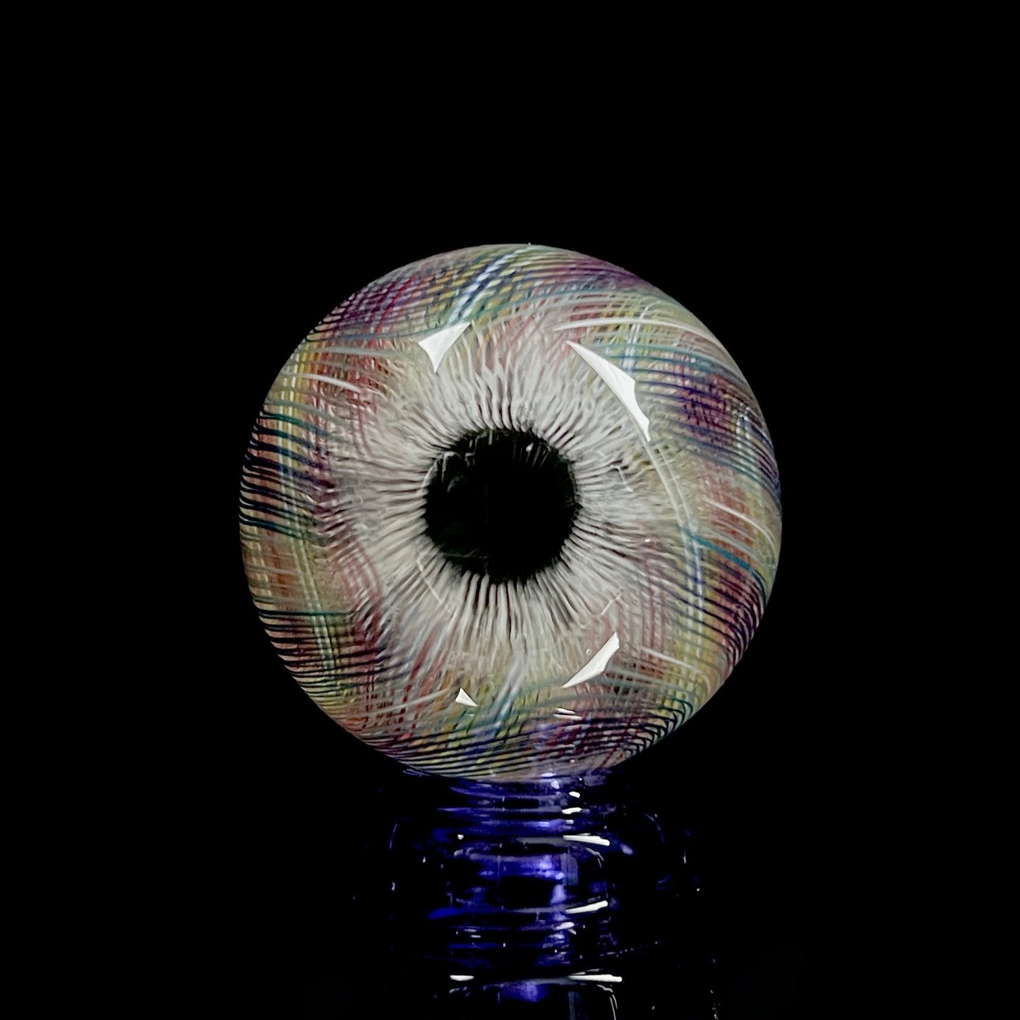 A multicolored glass marble with a mix of vibrant and muted tones, featuring a patterned design. The marble appears to have a convex surface.