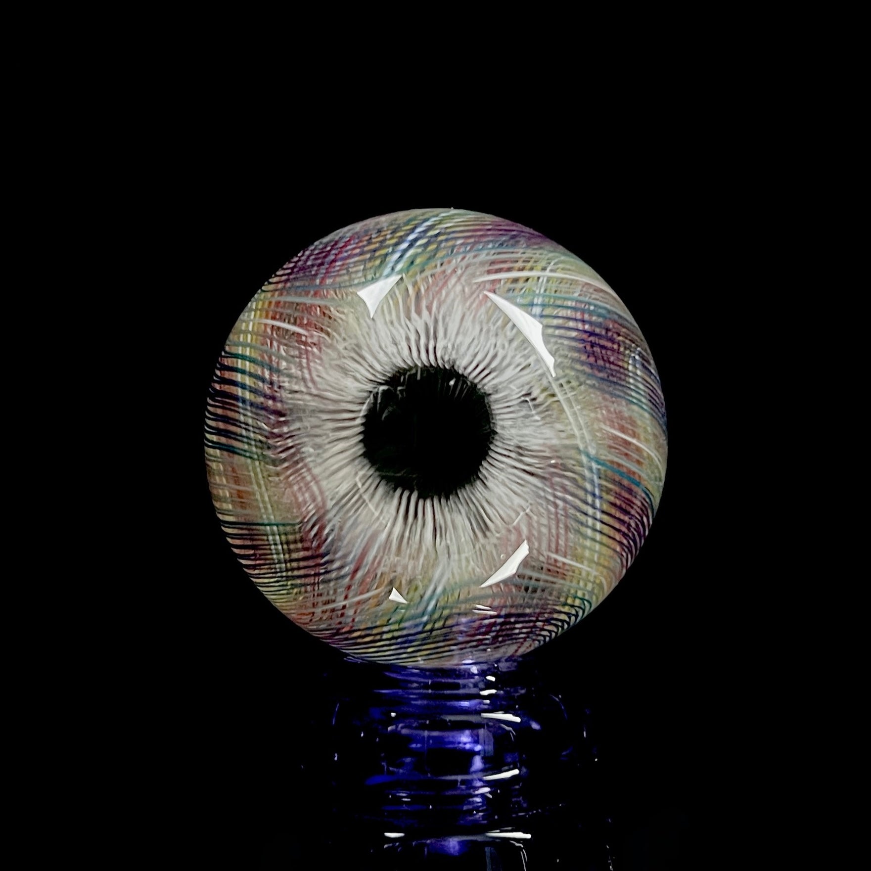 A multicolored glass marble with a mix of vibrant and muted tones, featuring a patterned design. The marble appears to have a convex surface.