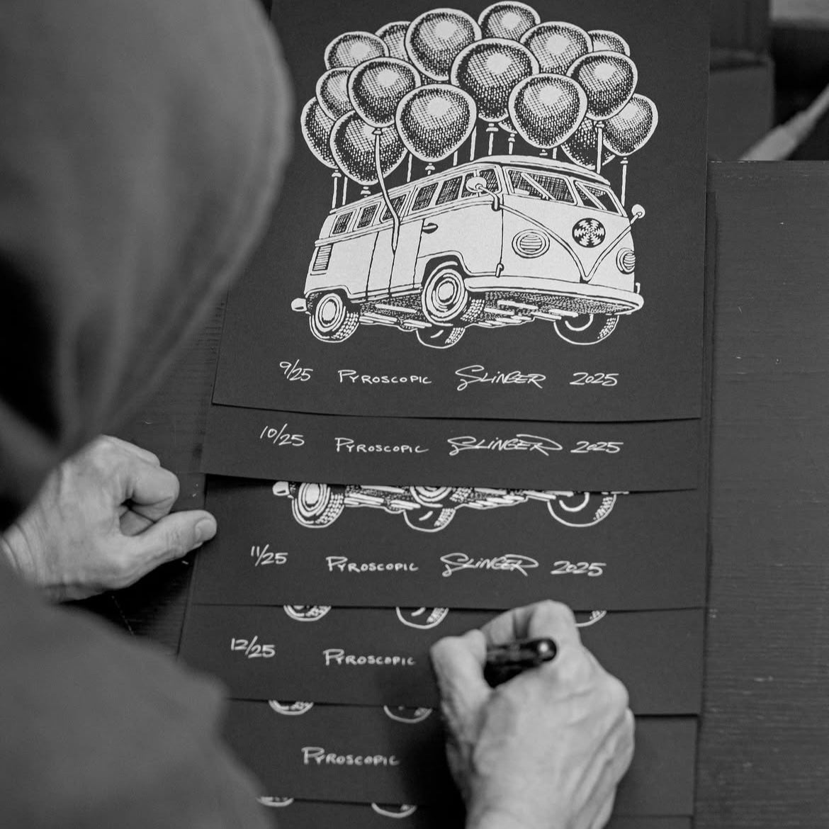 Person signing a stack of black and white prints featuring a vintage van with balloons.