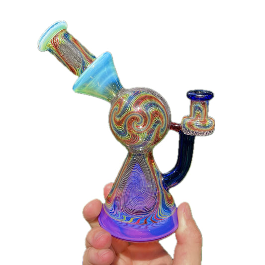 A multicolored glass piece with geometric patterns, featuring a small pyramid-shaped Perc and a narrow neck, held in a person's hand.