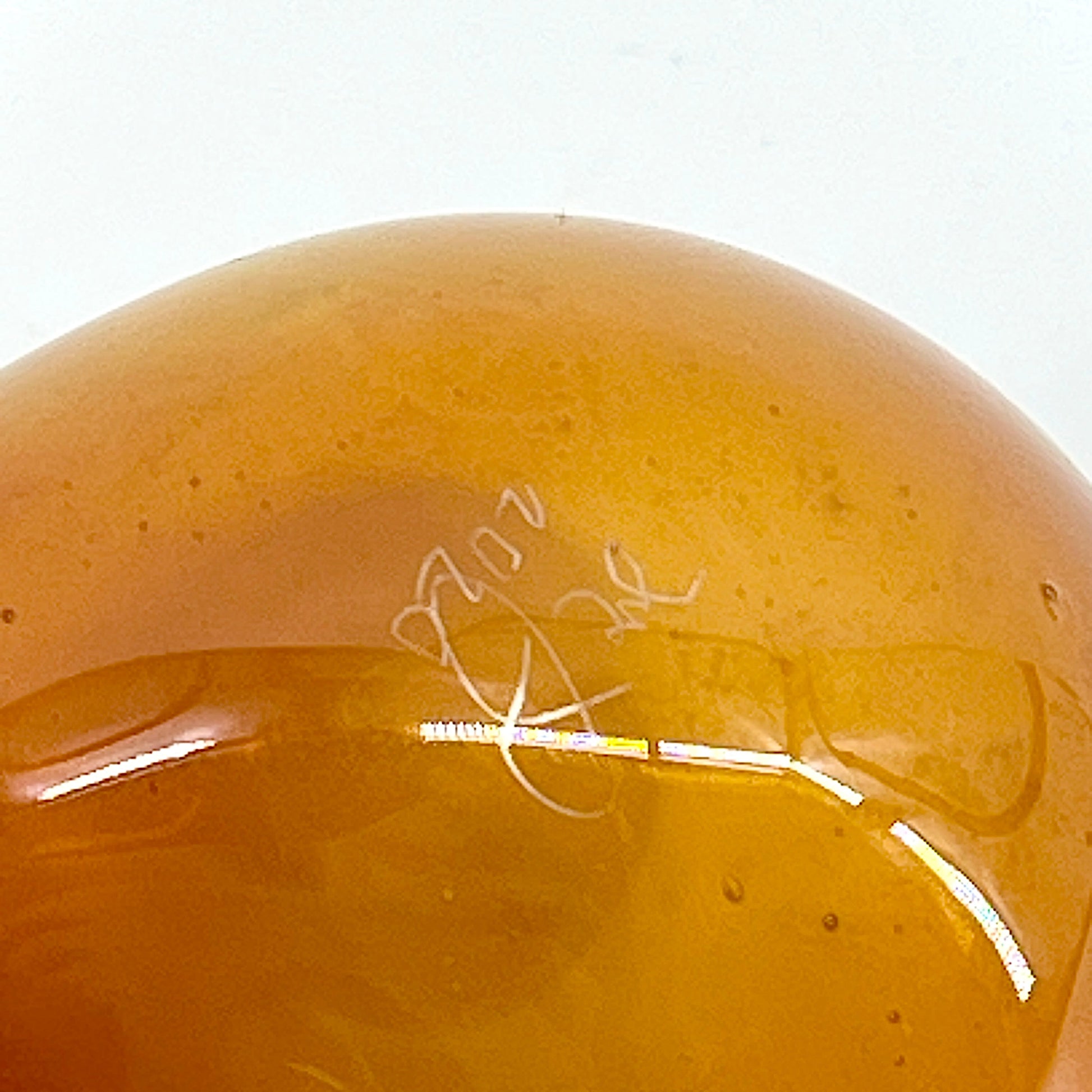Close-up of a yellowish-brown glass object with a signature on a white background.