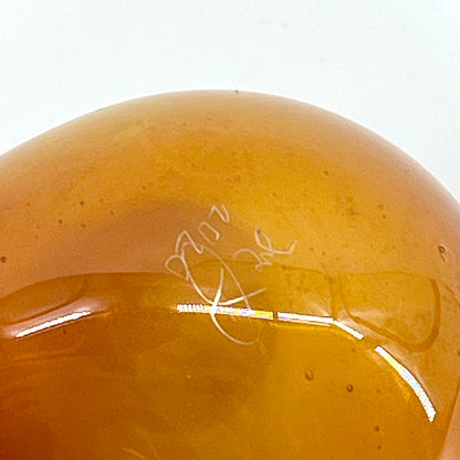 Close-up of a yellowish-brown glass object with a signature on a white background.