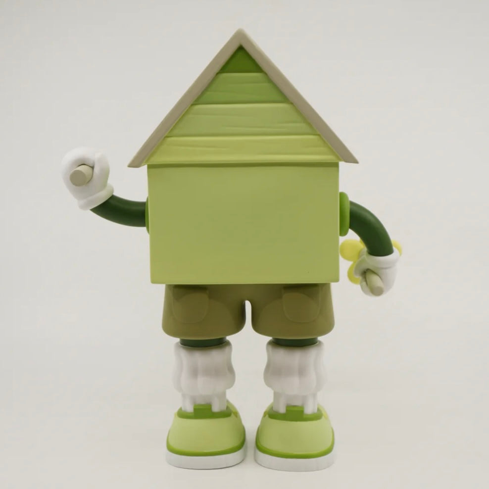 A toy figure of a character resembling a house with eyes, wearing green shorts, a green and white shirt, and green and white shoes, holding a yellow flower.