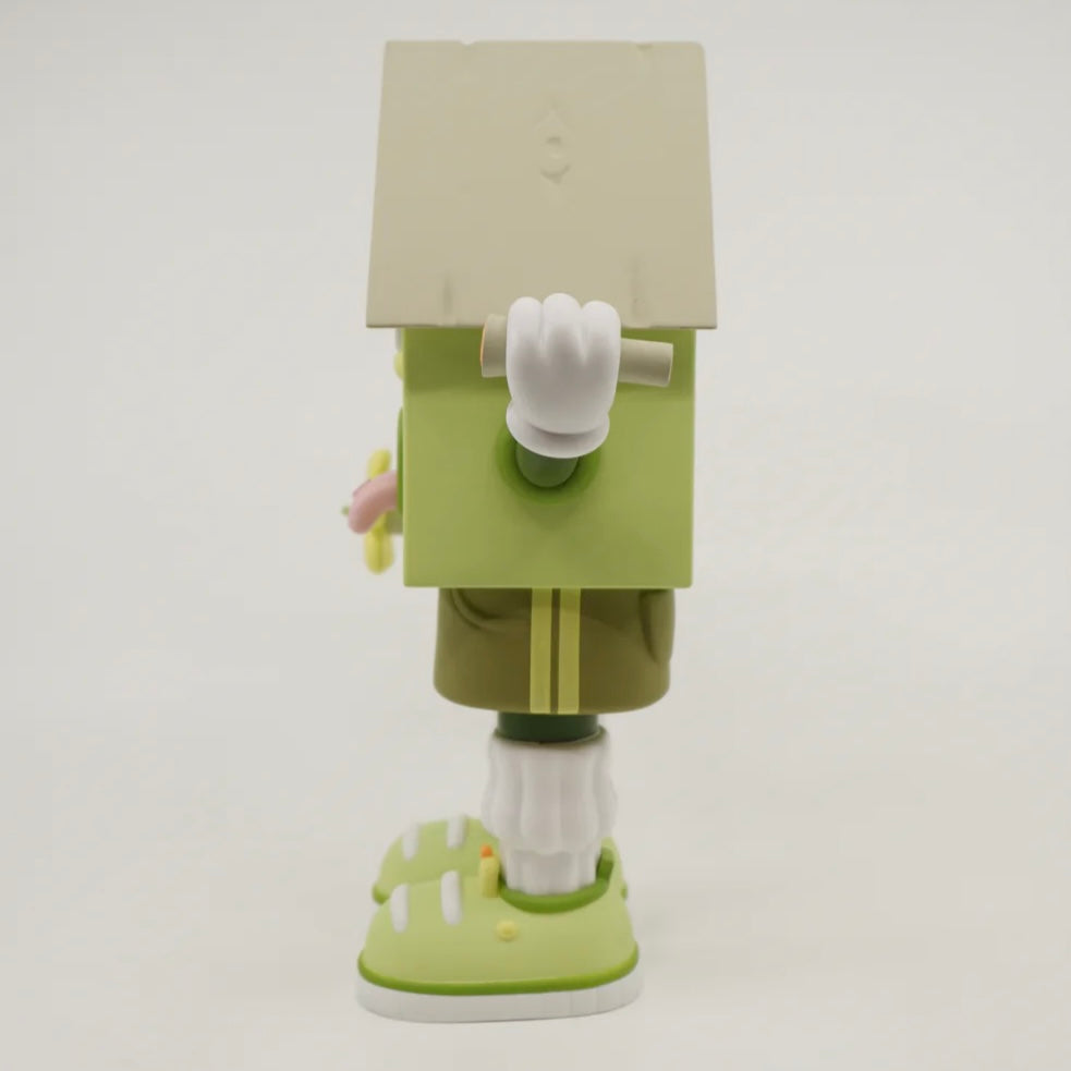 A toy figure of a character resembling a house with eyes, wearing green shorts, a green and white shirt, and green and white shoes, holding a yellow flower.
