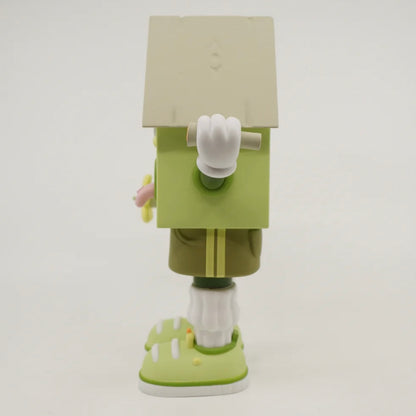 A toy figure of a character resembling a house with eyes, wearing green shorts, a green and white shirt, and green and white shoes, holding a yellow flower.
