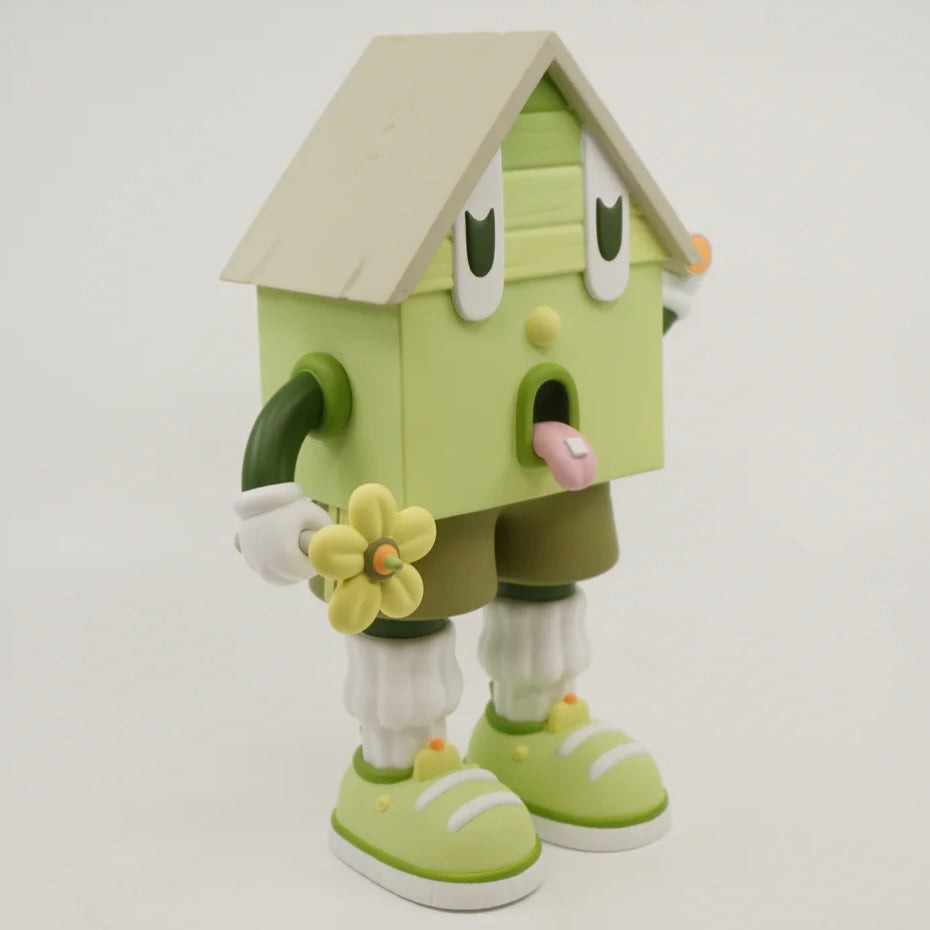 A toy figure of a character resembling a house with eyes, wearing green shorts, a green and white shirt, and green and white shoes, holding a yellow flower.