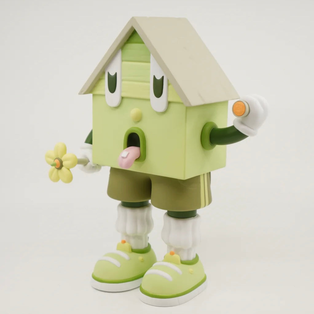 A toy figure of a character resembling a house with eyes, wearing green shorts, a green and white shirt, and green and white shoes, holding a yellow flower.