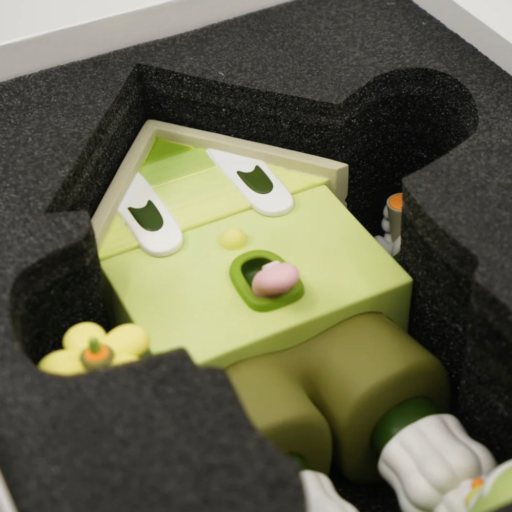 A toy figure of a character resembling a house with eyes, wearing green shorts, a green and white shirt, and green and white shoes, holding a yellow flower.