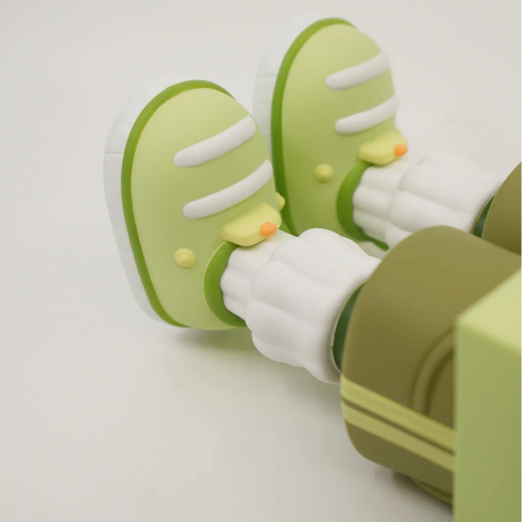 A toy figure of a character resembling a house with eyes, wearing green shorts, a green and white shirt, and green and white shoes, holding a yellow flower.