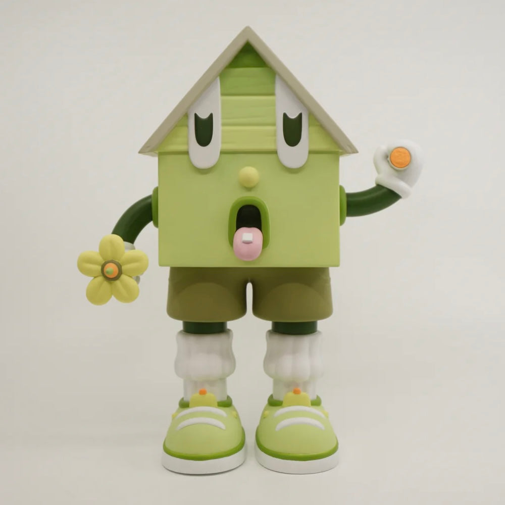 A toy figure of a character resembling a house with eyes, wearing green shorts, a green and white shirt, and green and white shoes, holding a yellow flower.