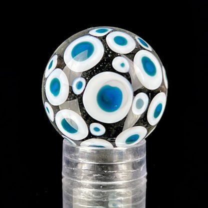 A glass marble with a design of blue dots on a white background, approximately 26mm in diameter, displayed on a clear stand.