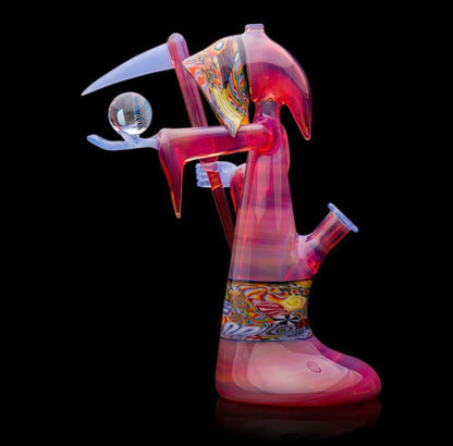 A colorful glass pipe shaped like a reaper with a scythe, featuring abstract designs and a removable dabber.