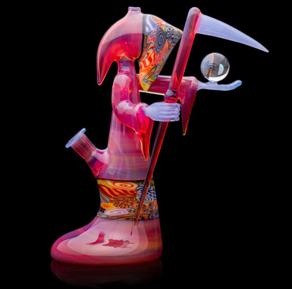A colorful glass pipe shaped like a reaper with a scythe, featuring abstract designs and a removable dabber.