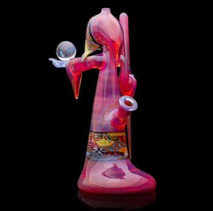 A colorful glass pipe shaped like a reaper with a scythe, featuring abstract designs and a removable dabber.