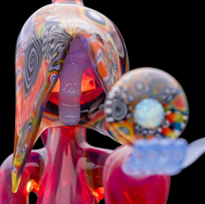 A colorful glass pipe shaped like a reaper with a scythe, featuring abstract designs and a removable dabber.